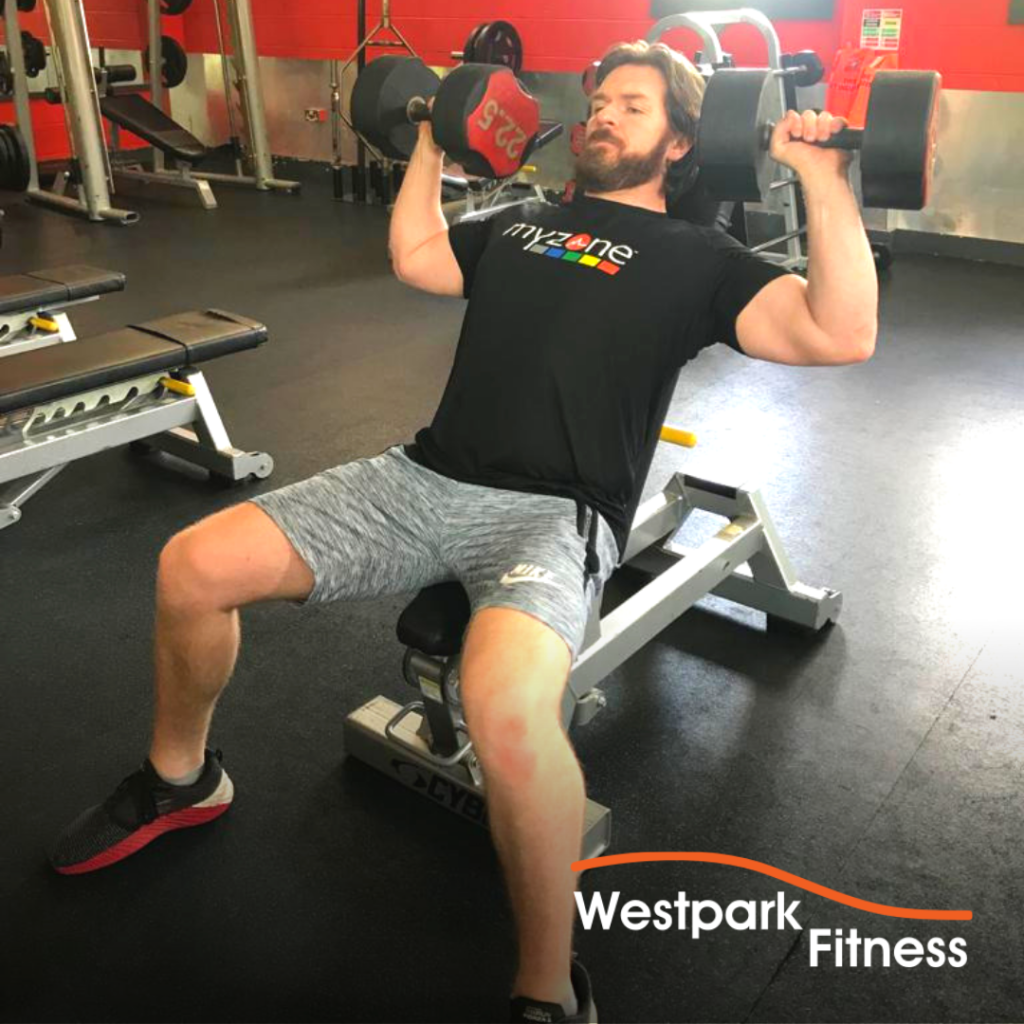 westpark fitness dumbbell shoulder press exercise male gym goer sitting on a bench holding a dumbbell at each side of his head