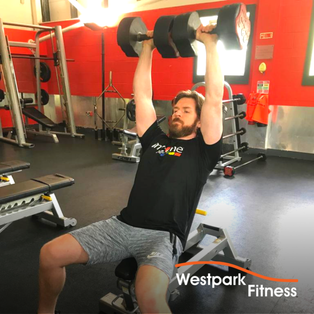 westpark fitness dumbbell shoulder press exercise male gym goer sitting on a bench holding two dumbbells over his head